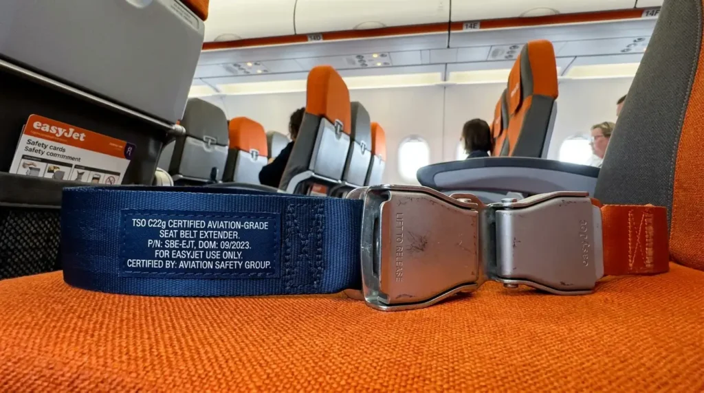 Certified aviation-grade seat belt extender with solid metal buckle and certification markings laid on an orange EasyJet aircraft seat alongside the standard aircraft lap belt